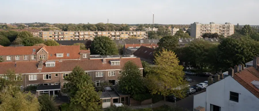 Overzicht van een woonwijk met rijtjeswoningen en naoorlogse bouw tussen de bomen, met een fabrieksschoorsteen aan de horizon